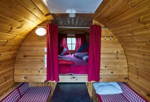 Campingfass von innen: Doppelbett hinter roten Vorhängen, seitlich zwei Sitzbänke